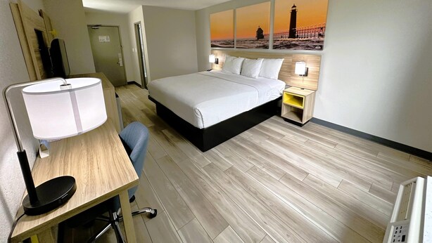 Habitación de hotel con cama, escritorio, silla, lámpara, mesita de noche y un cuadro de un faro.