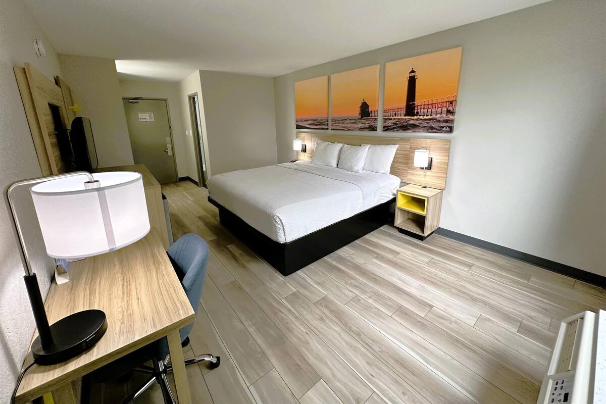A hotel room with a bed, desk, chair, lamp, nightstand, and a painting of a lighthouse.
