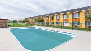 Una piscina con borde de azulejos, rodeada por una valla y un edificio con una sección de ladrillo a la izquierda.