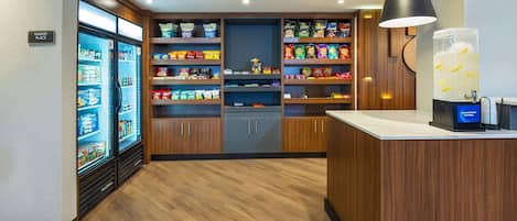A hotel room with a kitchenette, a refrigerator, shelves with snacks, a large water dispenser, and a sign indicating the market place.