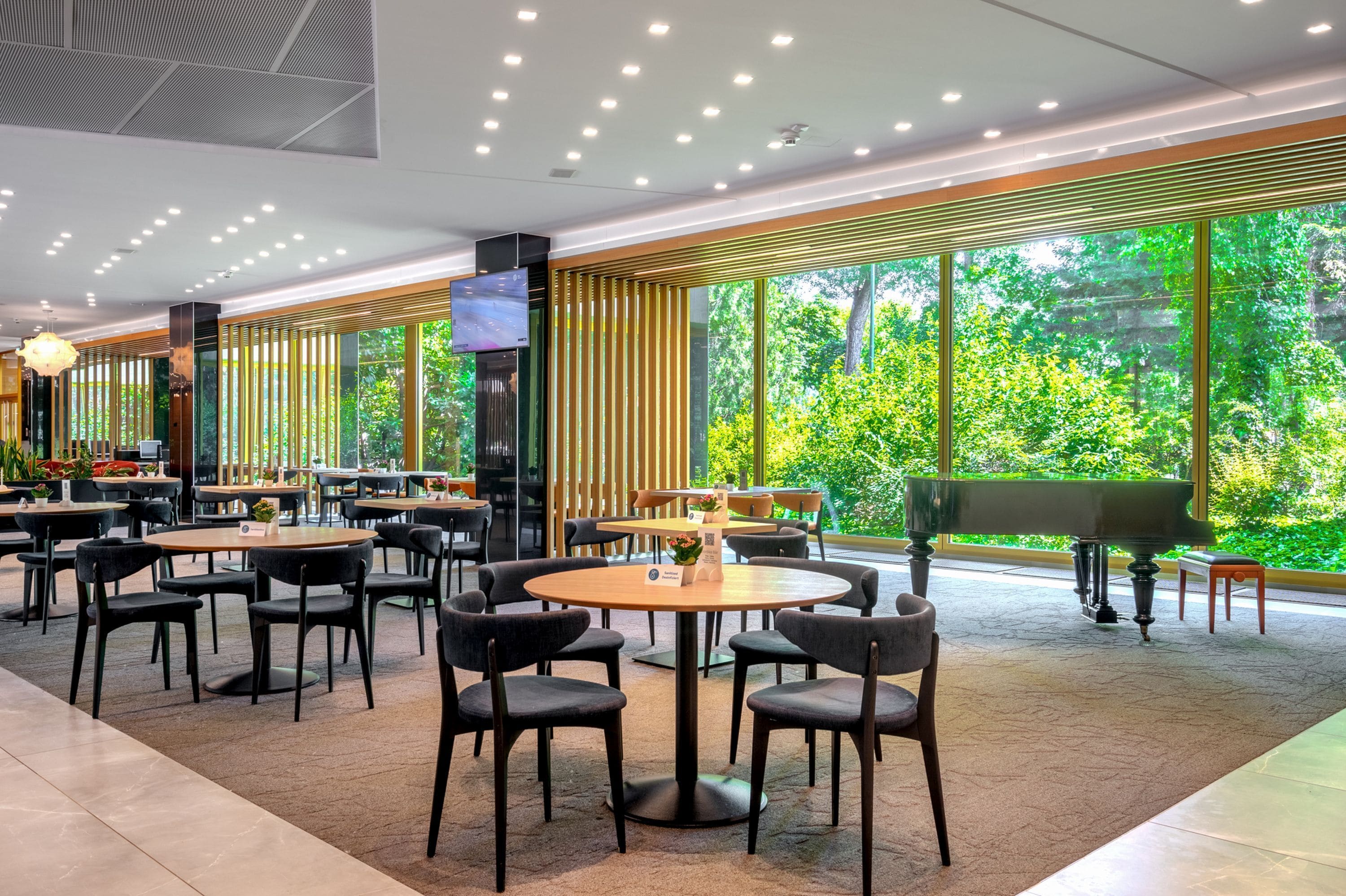 A modern indoor dining area with round tables and chairs, a grand piano, and large windows offering a view of greenery.