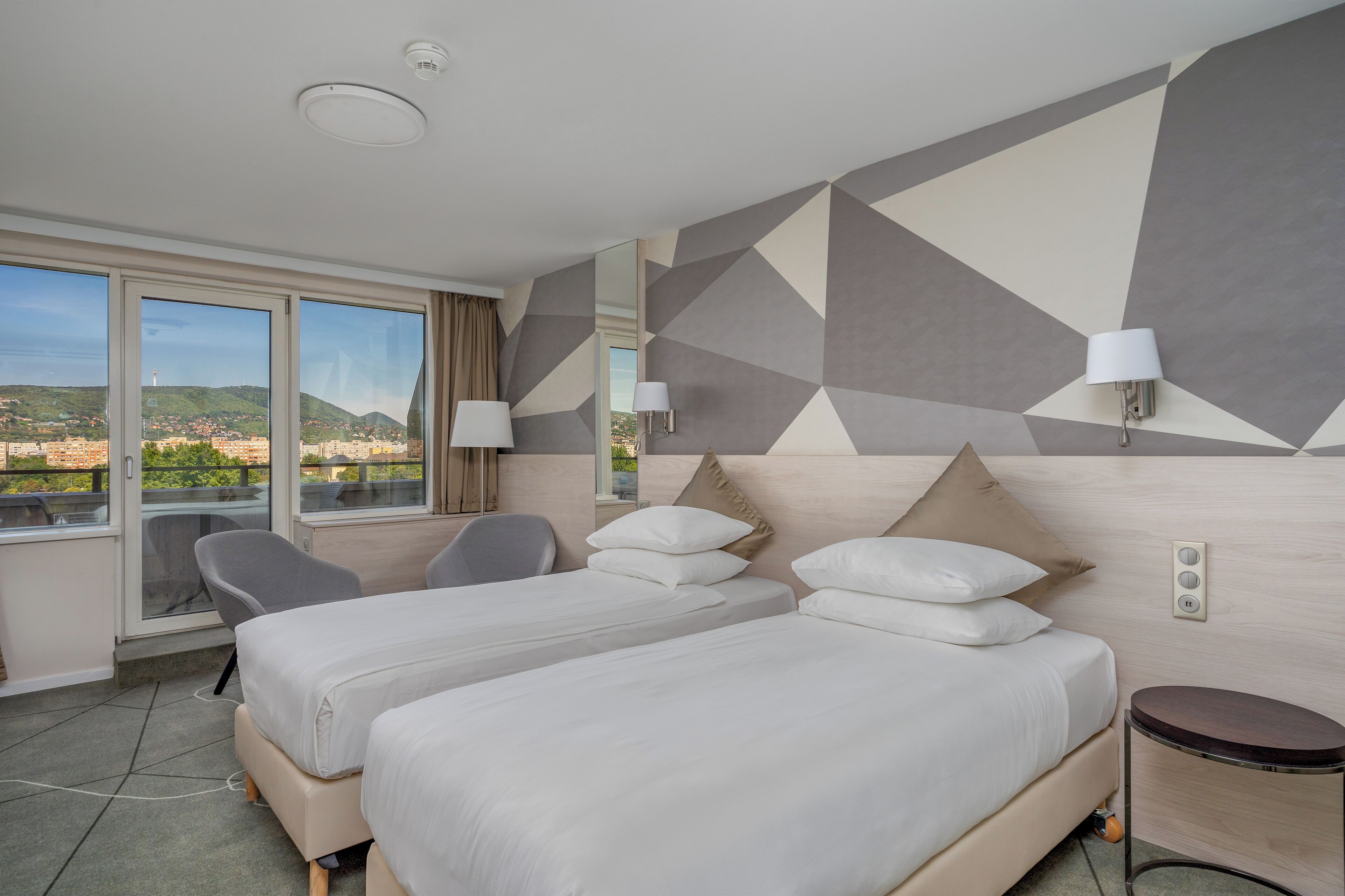 Habitación de hotel con dos camas, una silla, una mesita y vista a la ciudad.