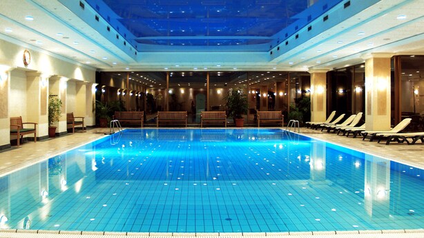 An indoor swimming pool with a tiled floor, wooden benches, and potted plants. - Ensana Thermal Margaret Island (Budapest)