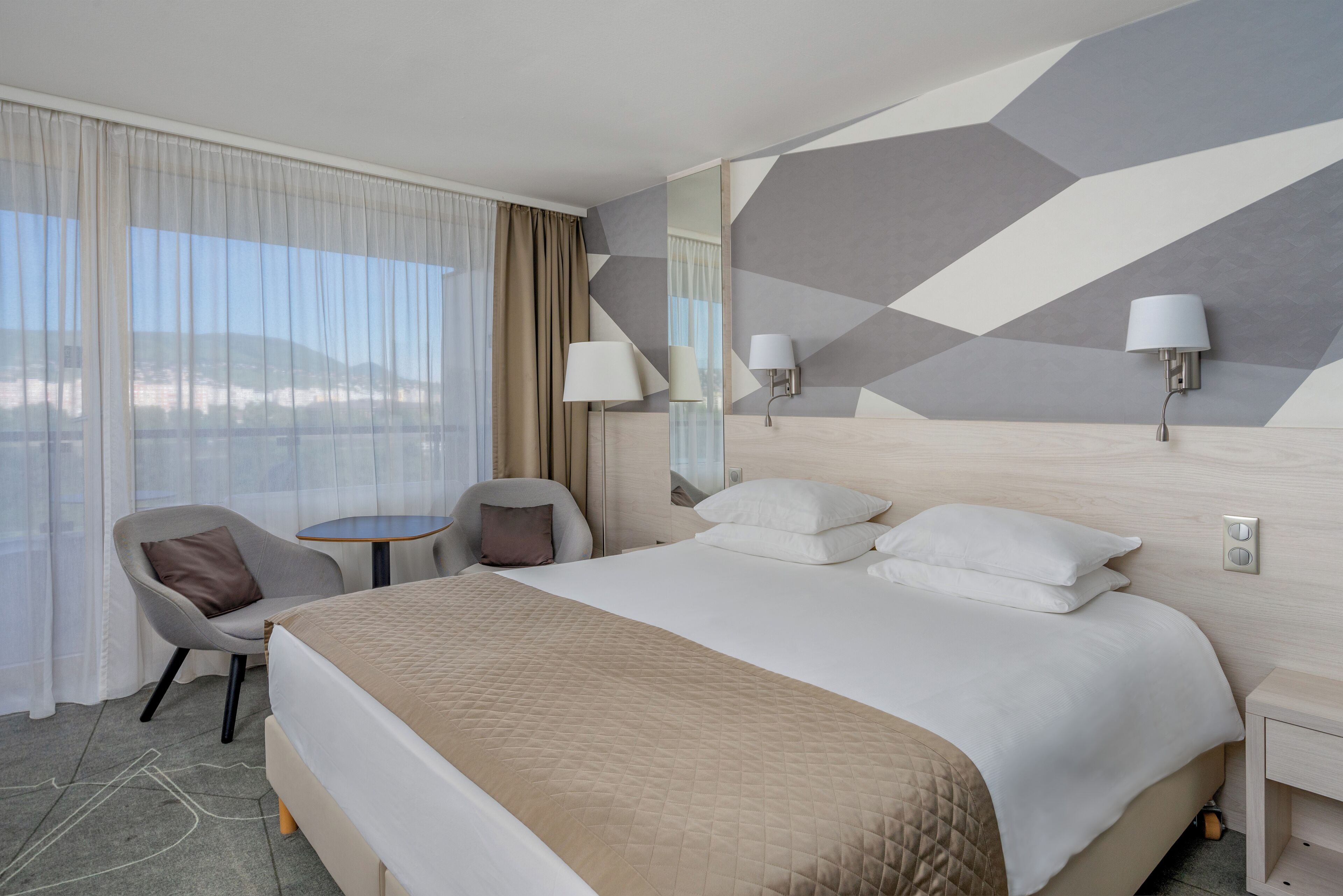 A hotel room with a large bed, a chair, a small table, and a view of the mountains.