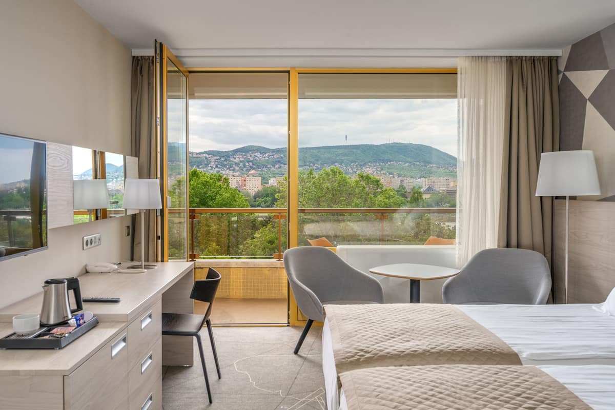 Ein Hotelzimmer mit Bett, Schreibtisch, Stuhl, Tisch und einem Balkon mit Blick auf die Stadt und die Hügel.