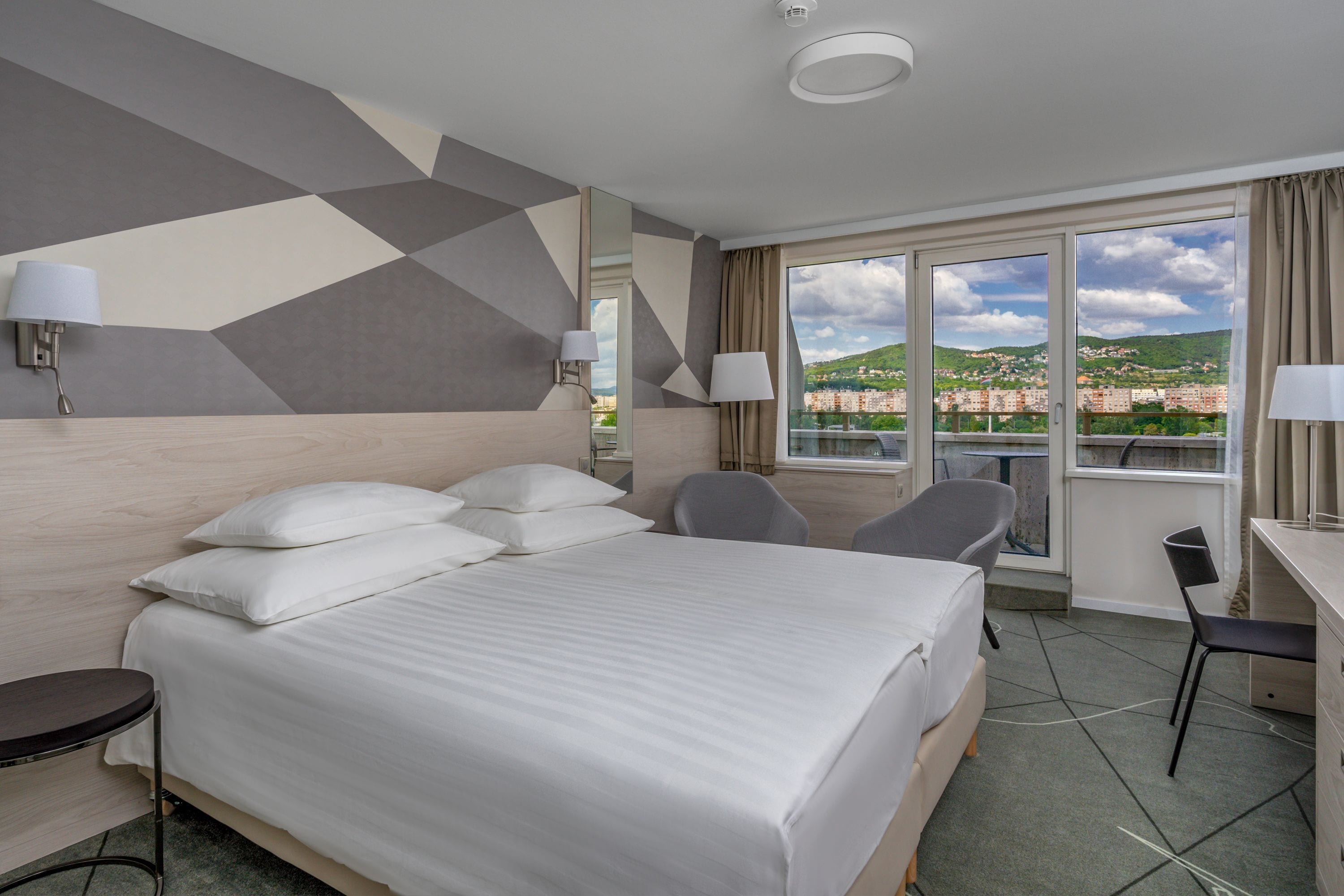 A hotel room with a large bed, two chairs, a desk, and a view of a cityscape through the windows.