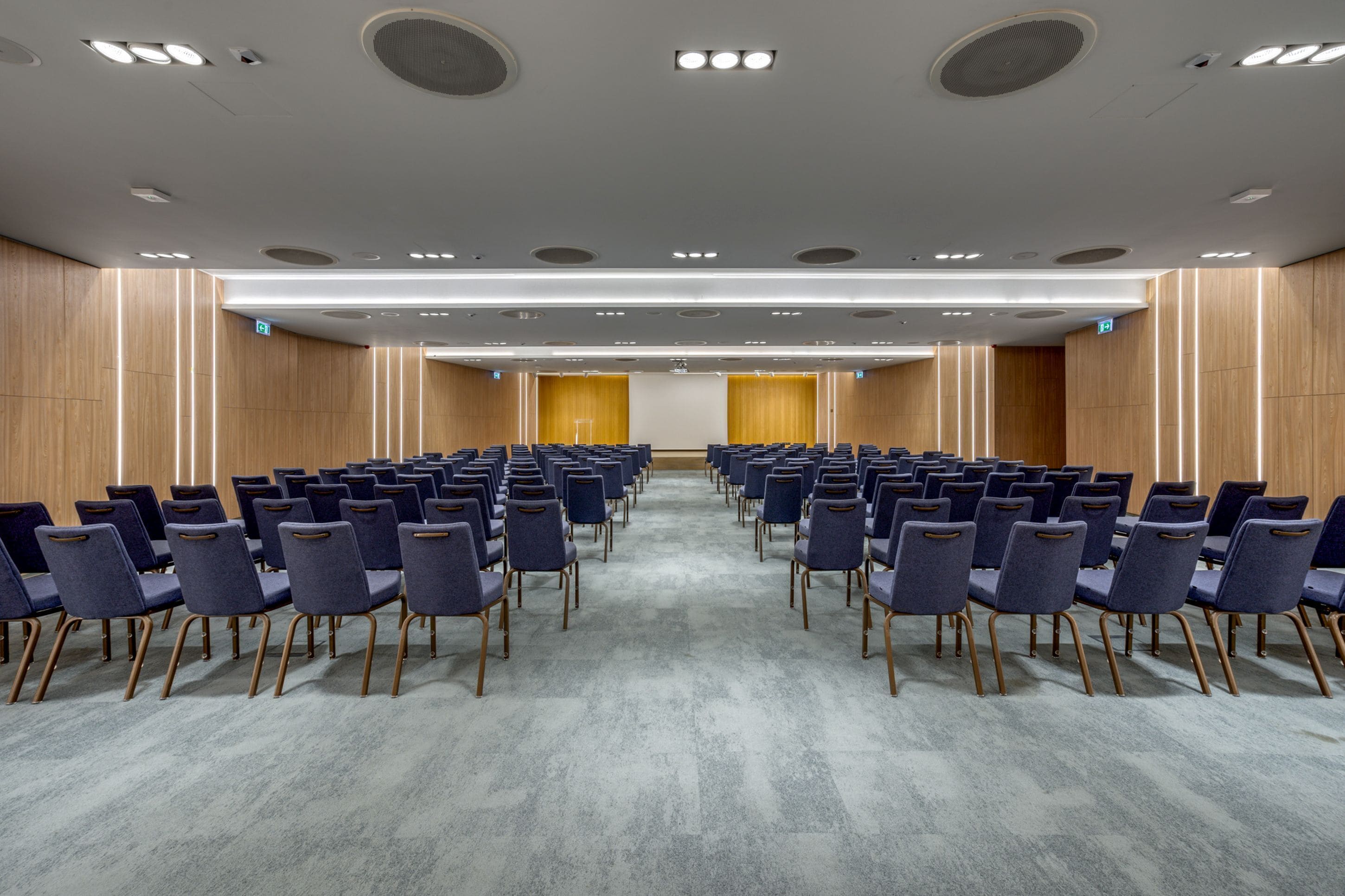 A conference room with rows of chairs, a carpeted floor, wooden panel walls, and recessed ceiling lights.