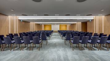 A conference room with rows of chairs, a carpeted floor, wooden panel walls, and recessed ceiling lights.