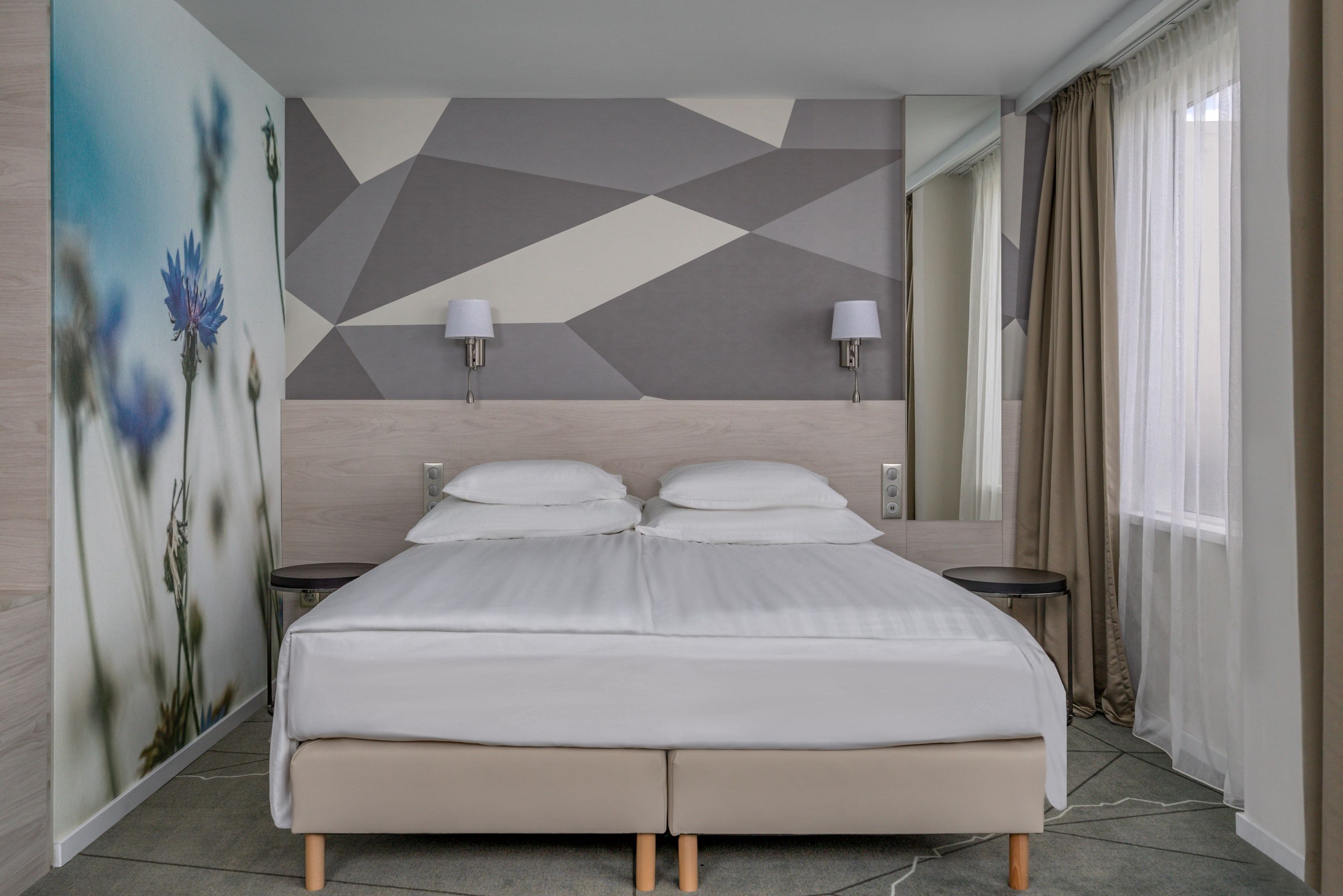 Una habitación de hotel moderna con una cama grande, dos mesitas de noche, un muro con estampado geométrico y un amplio ventanal con cortinas.