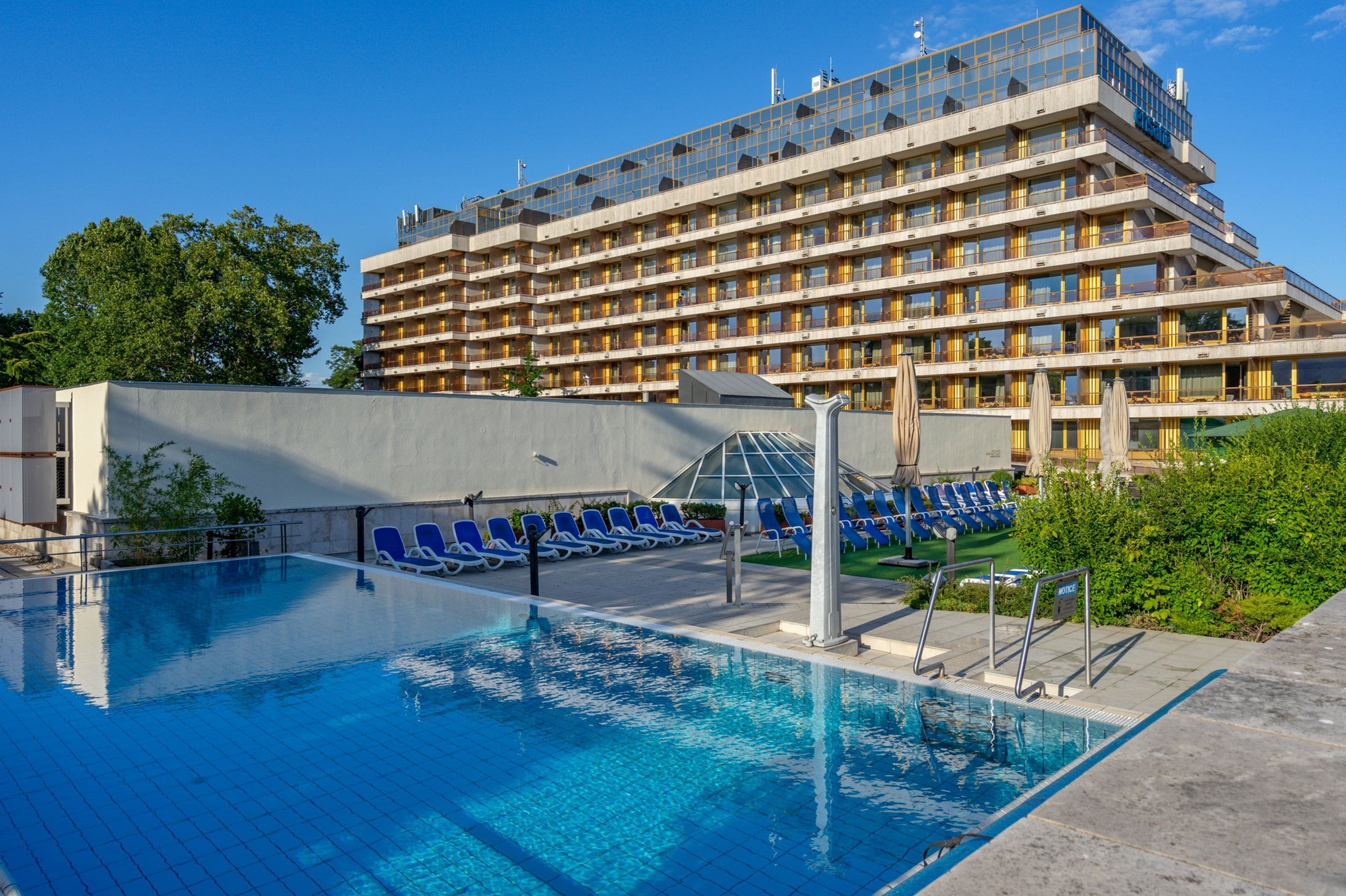 Un hôtel doté d’une piscine, de chaises longues bleues et d’un grand bâtiment à plusieurs étages et balcons.