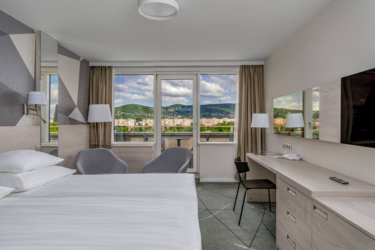 Ein Hotelzimmer mit Bett, zwei Stühlen, einem Schreibtisch, einem Spiegel und Blick auf eine Stadtlandschaft.