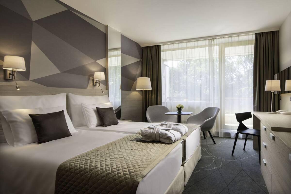 Ein Hotelzimmer mit einem großen Bett, zwei Nachttischen, einem Schreibtisch, zwei Stühlen und einem Fenster mit Vorhängen.