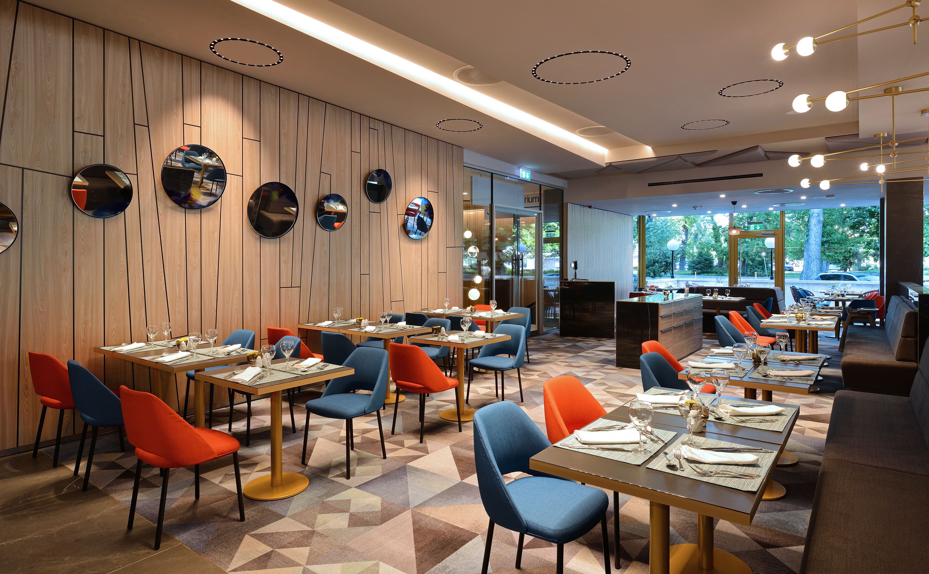Interior de restaurante com paredes de painel de madeira, espelhos redondos e luminárias modernas. Mesas com cadeiras azuis e laranjas, e o chão com design de azulejos geométricos.