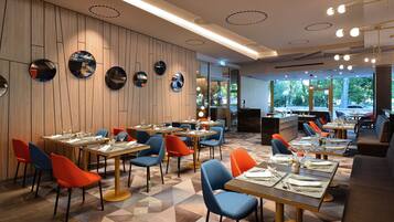 Interior de restaurante com paredes de painel de madeira, espelhos redondos e luminárias modernas. Mesas com cadeiras azuis e laranjas, e o chão com design de azulejos geométricos.