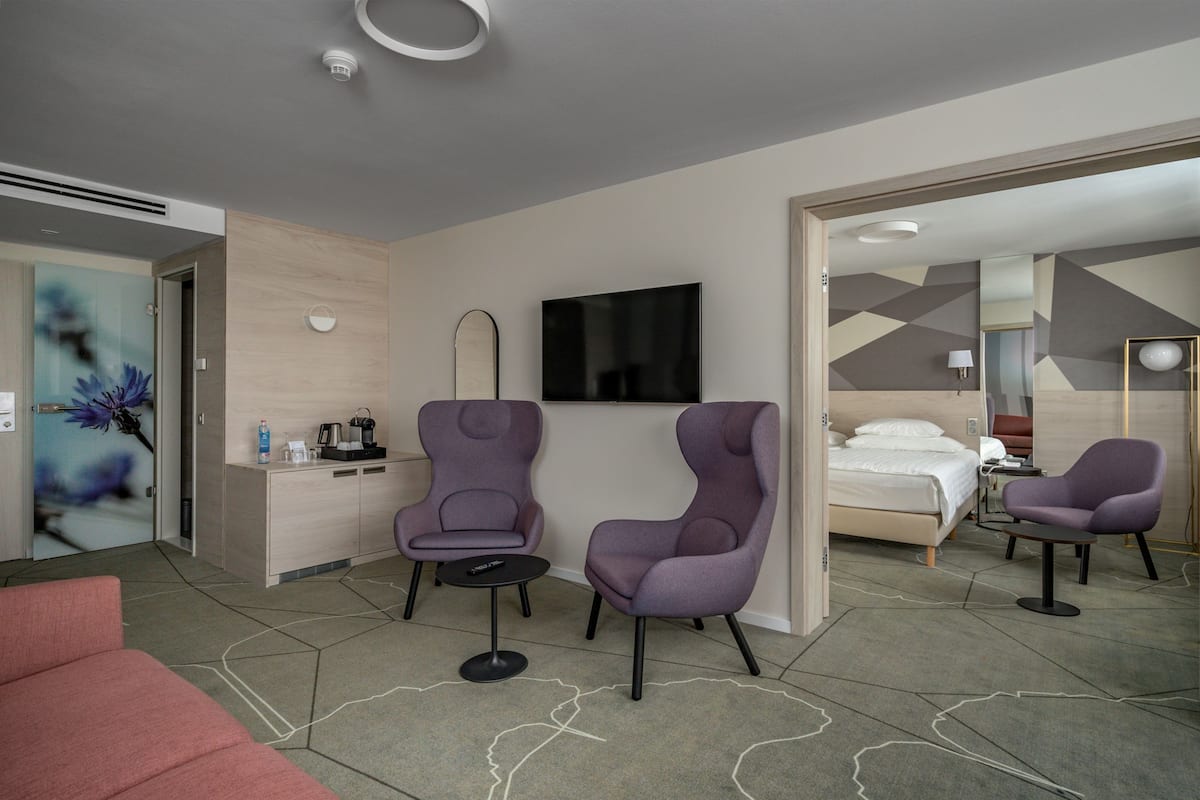 Ein Hotelzimmer mit einem Bett, zwei violetten Sessel, einem kleinen Tisch, einem Fernseher und einer Minibar.