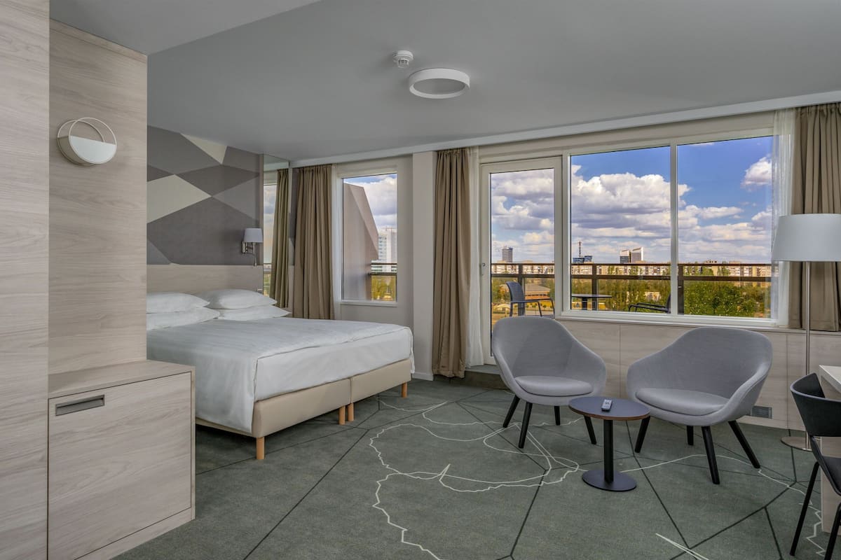 Ein modernes Hotelzimmer mit einem Bett, zwei Stühlen, einem kleinen Tisch und Blick auf die Stadt durch große Fenster.