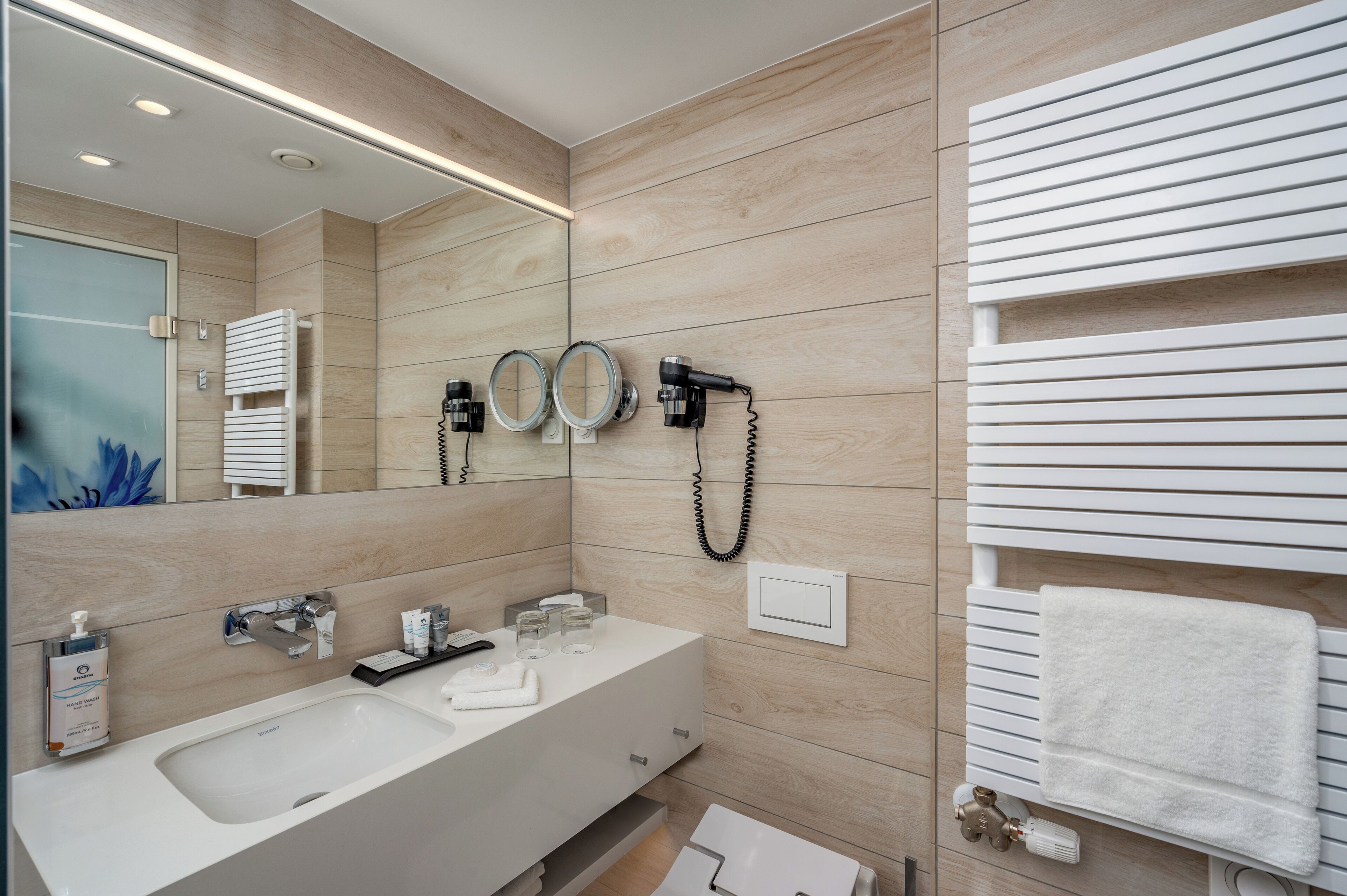 Un baño moderno con un espejo grande, dos secadores de pelo, un perchero para toallas y un lavamanos con dispensador de jabón.