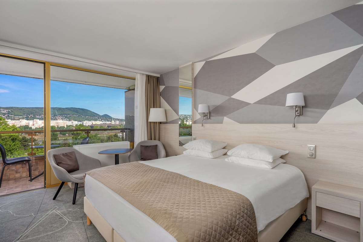Ein Hotelzimmer mit einem großen Bett, einem Schreibtisch und einem Sessel. Ein Balkon bietet Blick auf die Stadt und die Berge.
