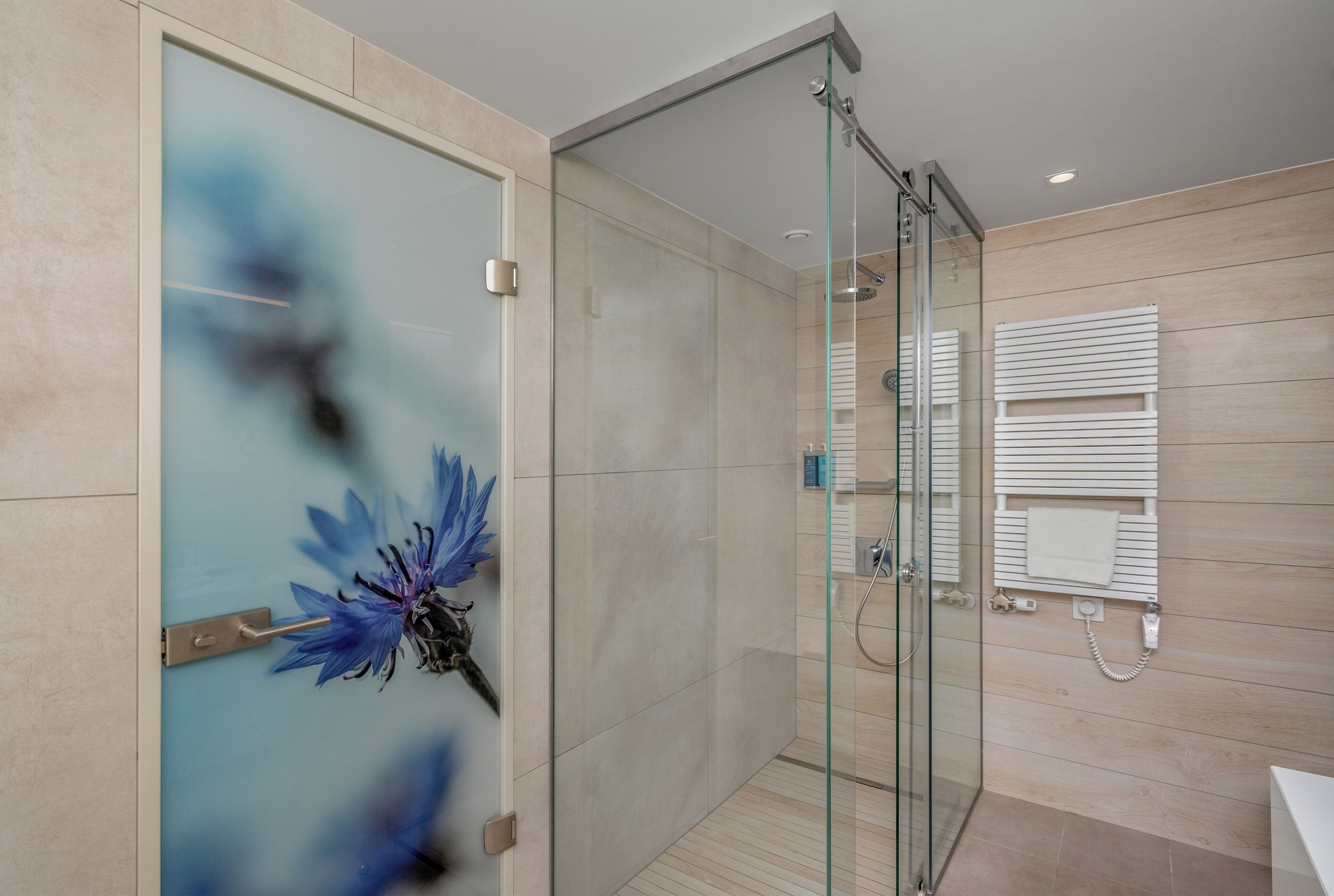 Un baño moderno con cabina de ducha de vidrio, suelo de madera y un diseño floral azul decorativo en la puerta.