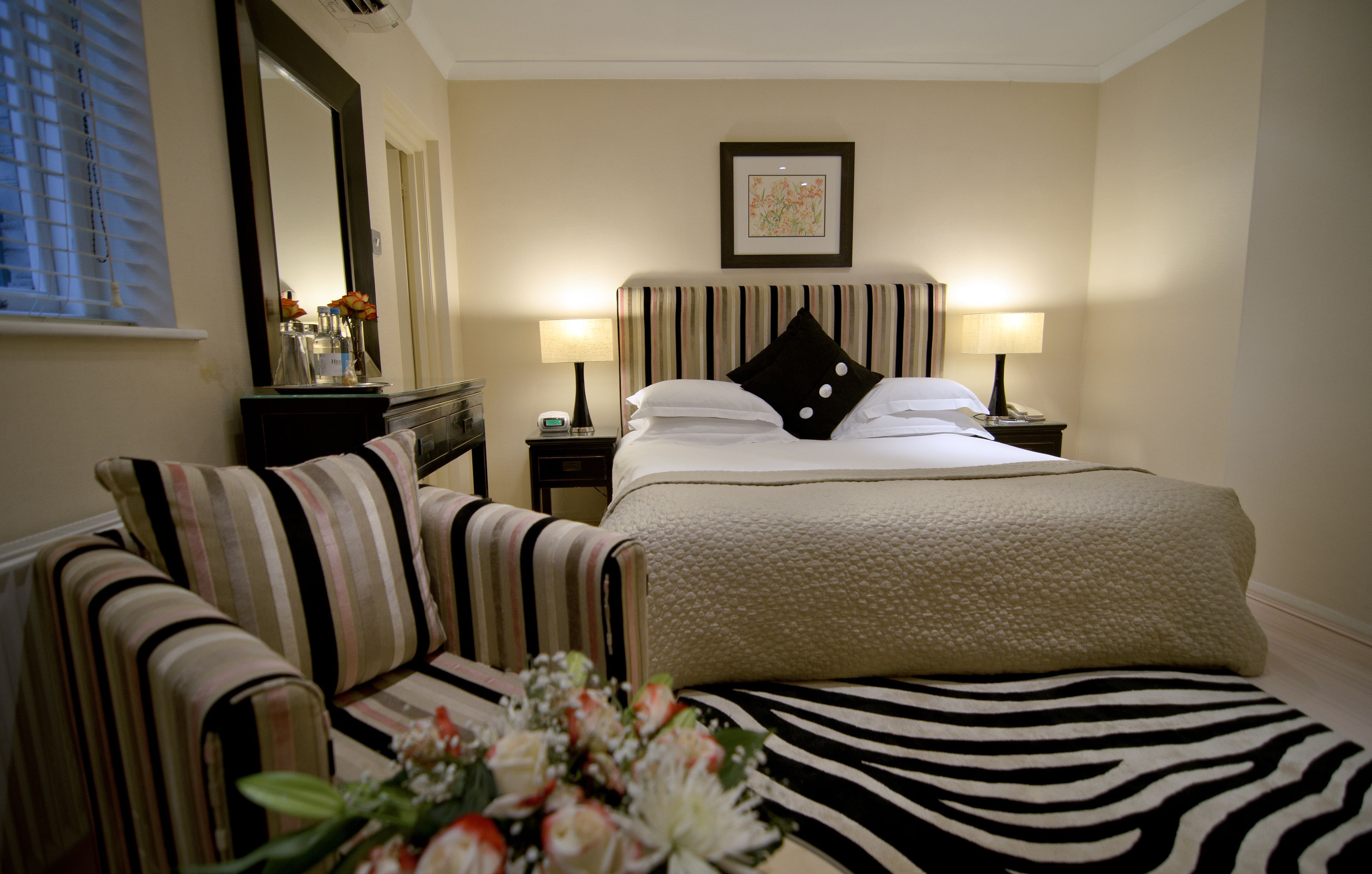 A hotel room with a bed, two bedside tables with lamps, a chair, a zebra-patterned rug, and a mirror on the wall.