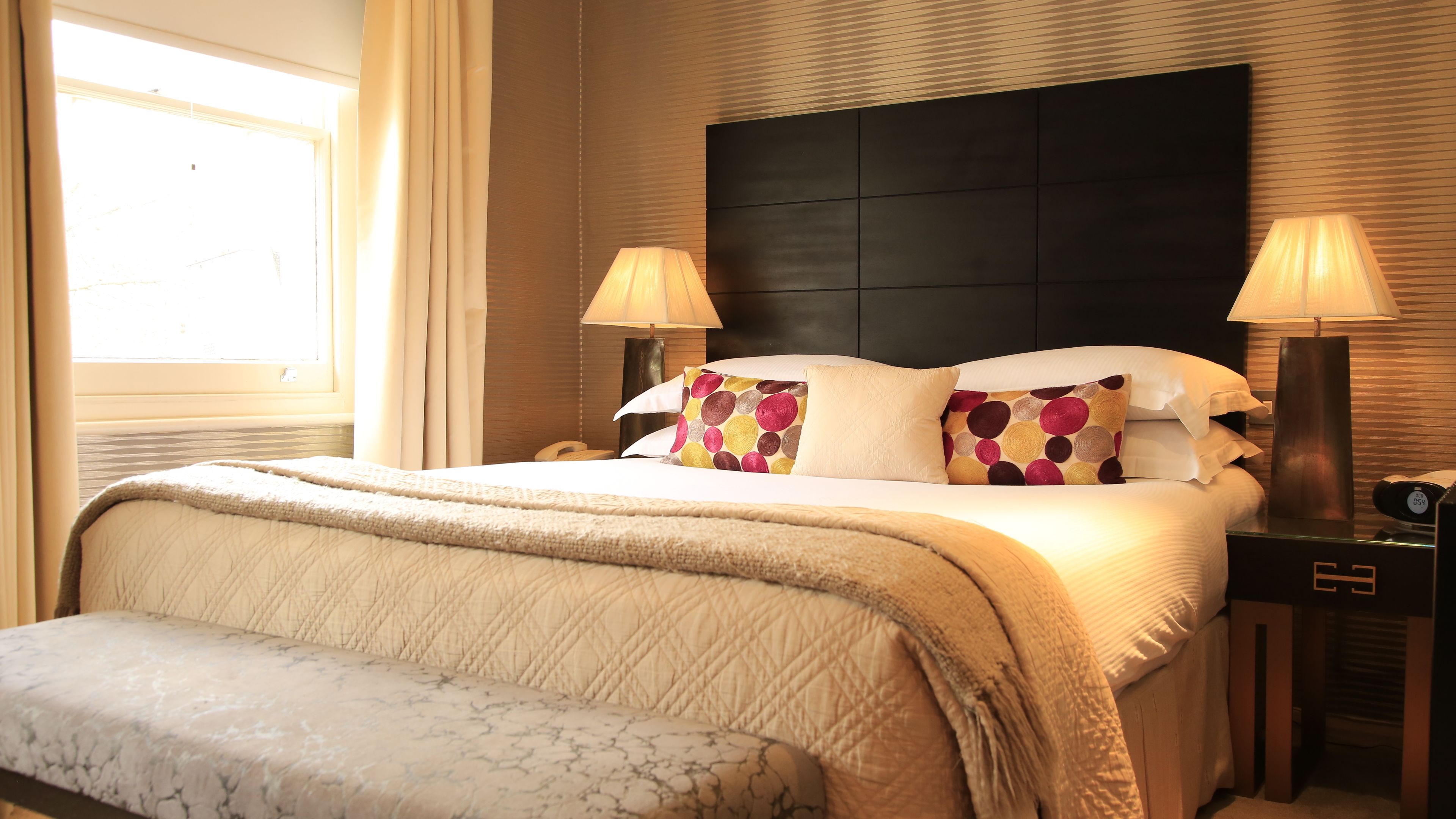 A hotel room with a large bed, two bedside lamps, a bedside clock, and a window with curtains.