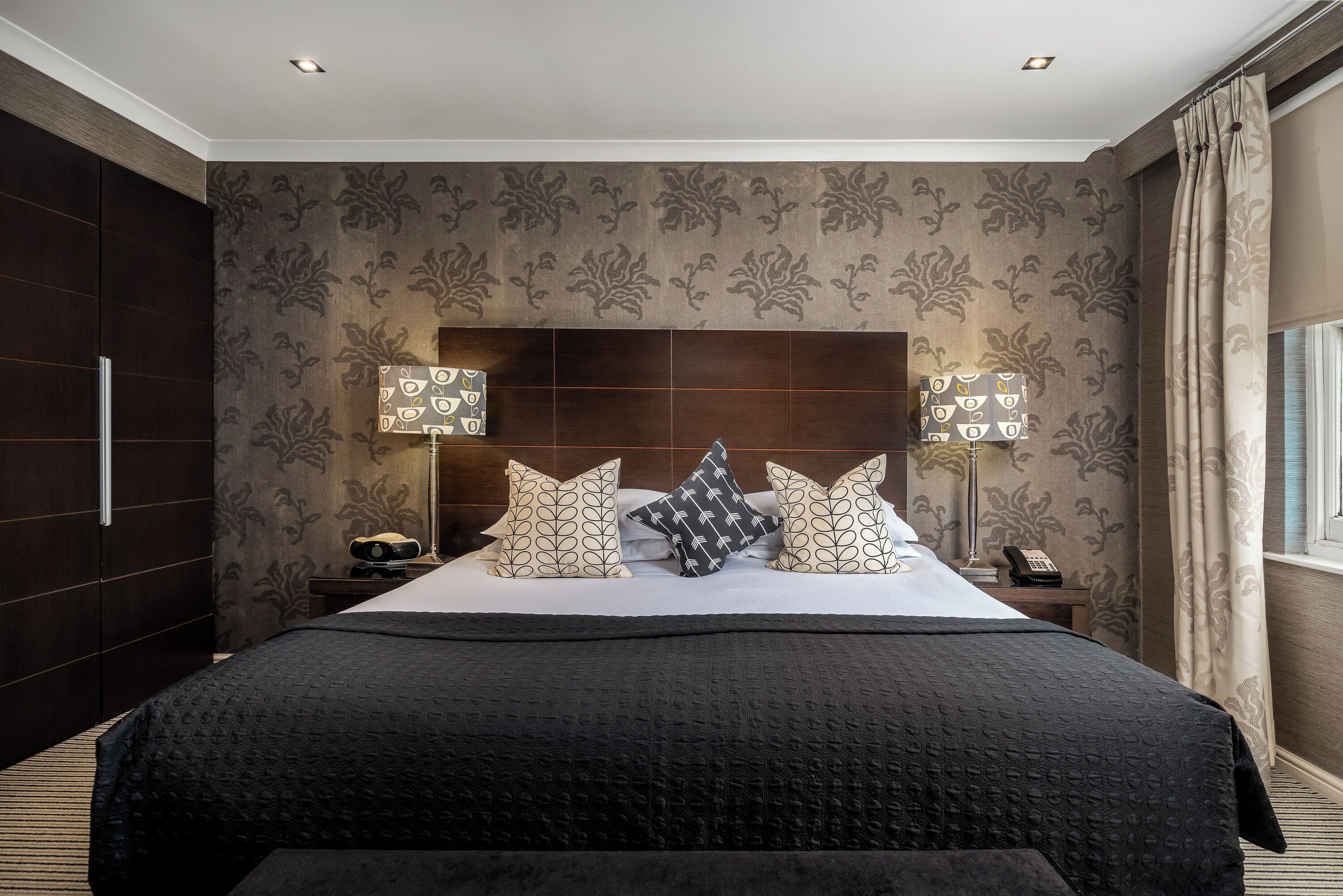 A hotel room with a large bed, dark wood headboard, patterned wallpaper, two bedside tables with lamps, and a window with curtains.