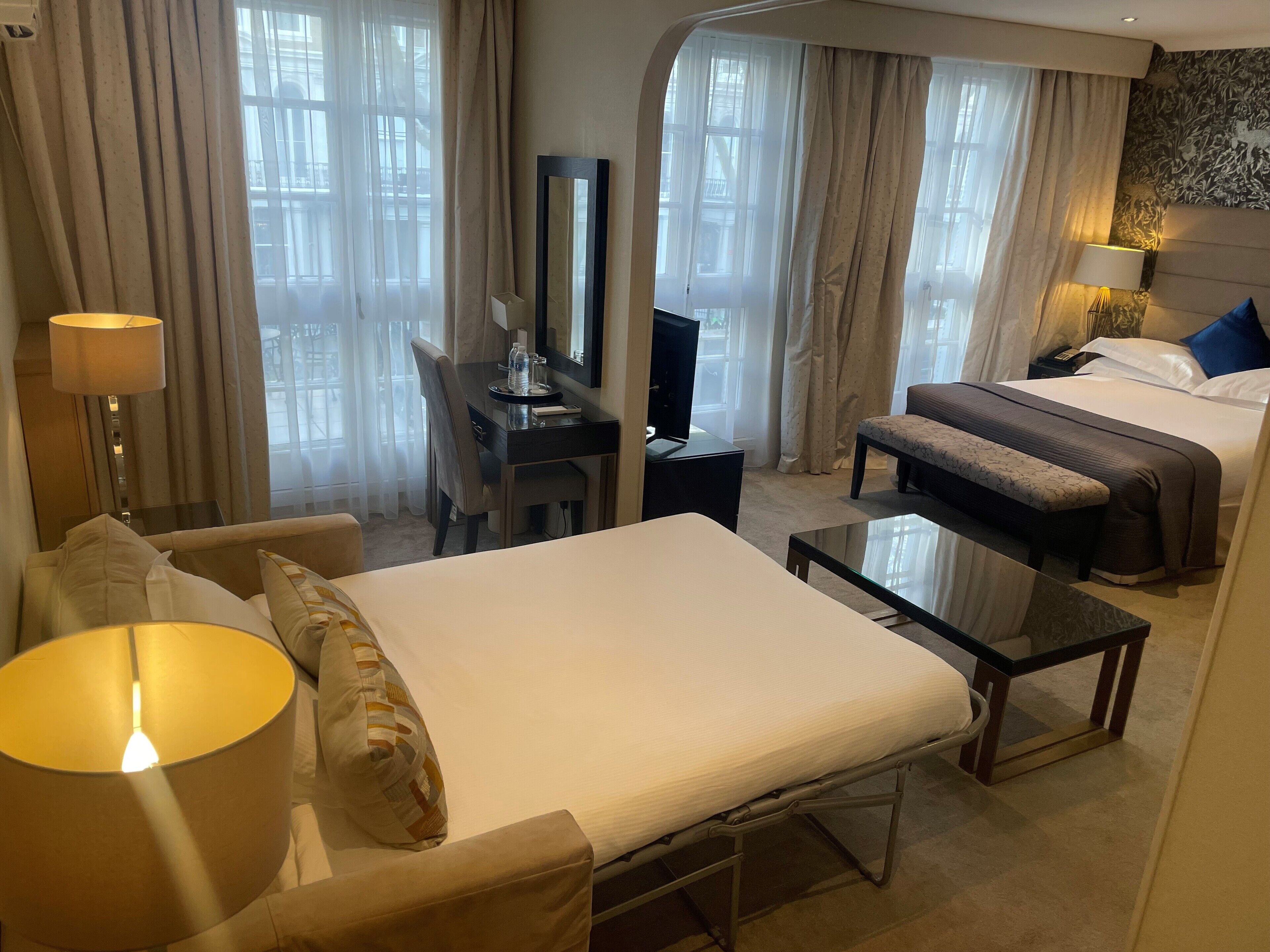 A hotel room with a bed, a desk, a chair, a small table, and a lamp.