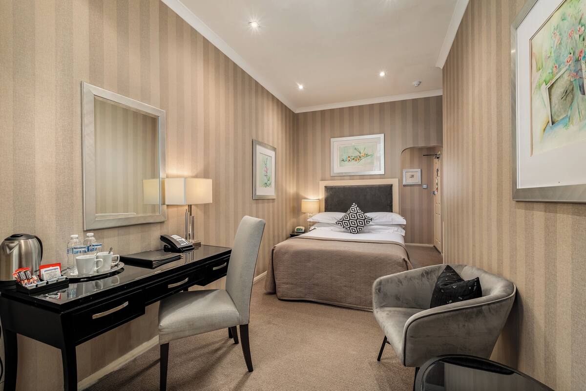 A hotel room with a bed, a desk with a chair, a bedside table with a lamp, a coffee maker, and a framed picture on the wall.