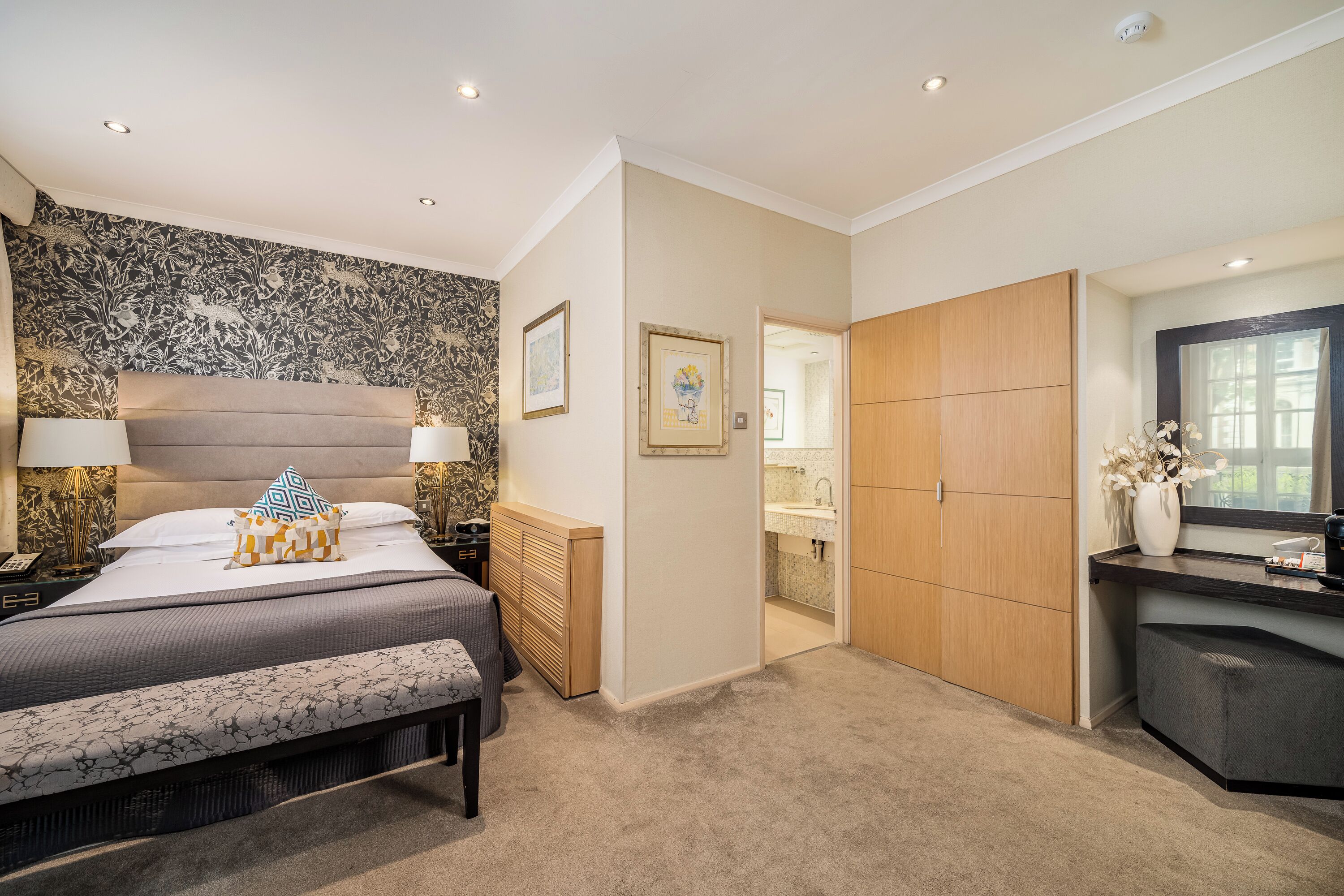 A modern bedroom with a large bed, bedside lamps, a bench, a wooden wardrobe, and a bathroom visible through an open door.