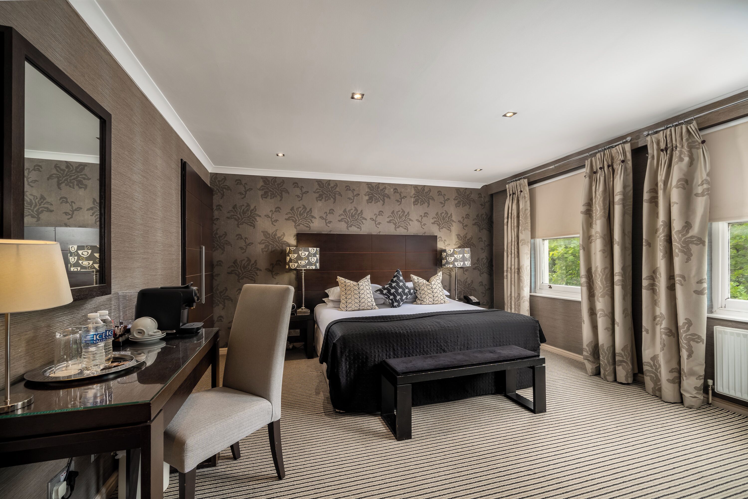 A hotel room with a bed, bedside tables, a desk, a chair, a bench, and a large window with curtains.