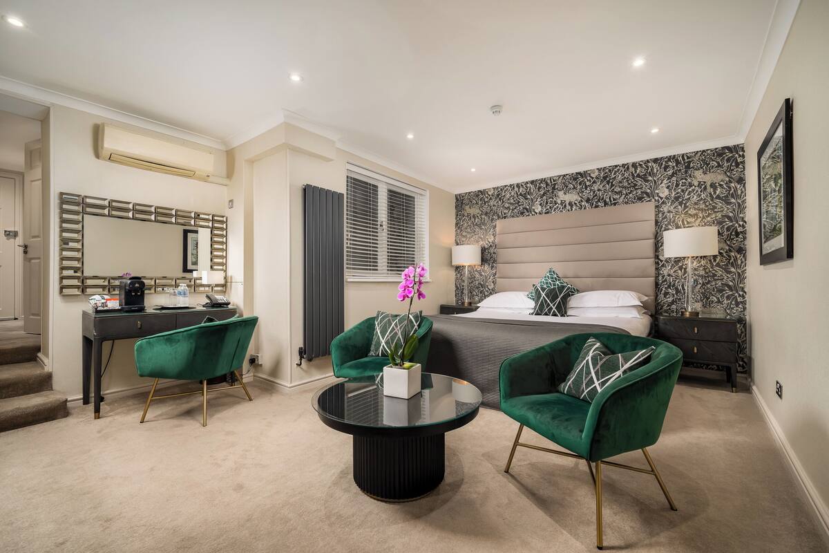 A modern hotel room with a bed, two green armchairs, a round coffee table, a desk with a computer, and a large mirror.