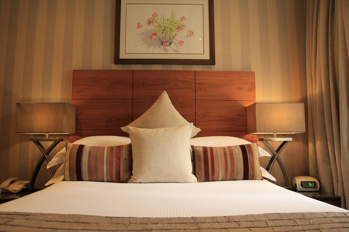 A hotel room with a wooden headboard, two bedside lamps, a telephone, a digital clock, and a framed floral artwork on the wall.