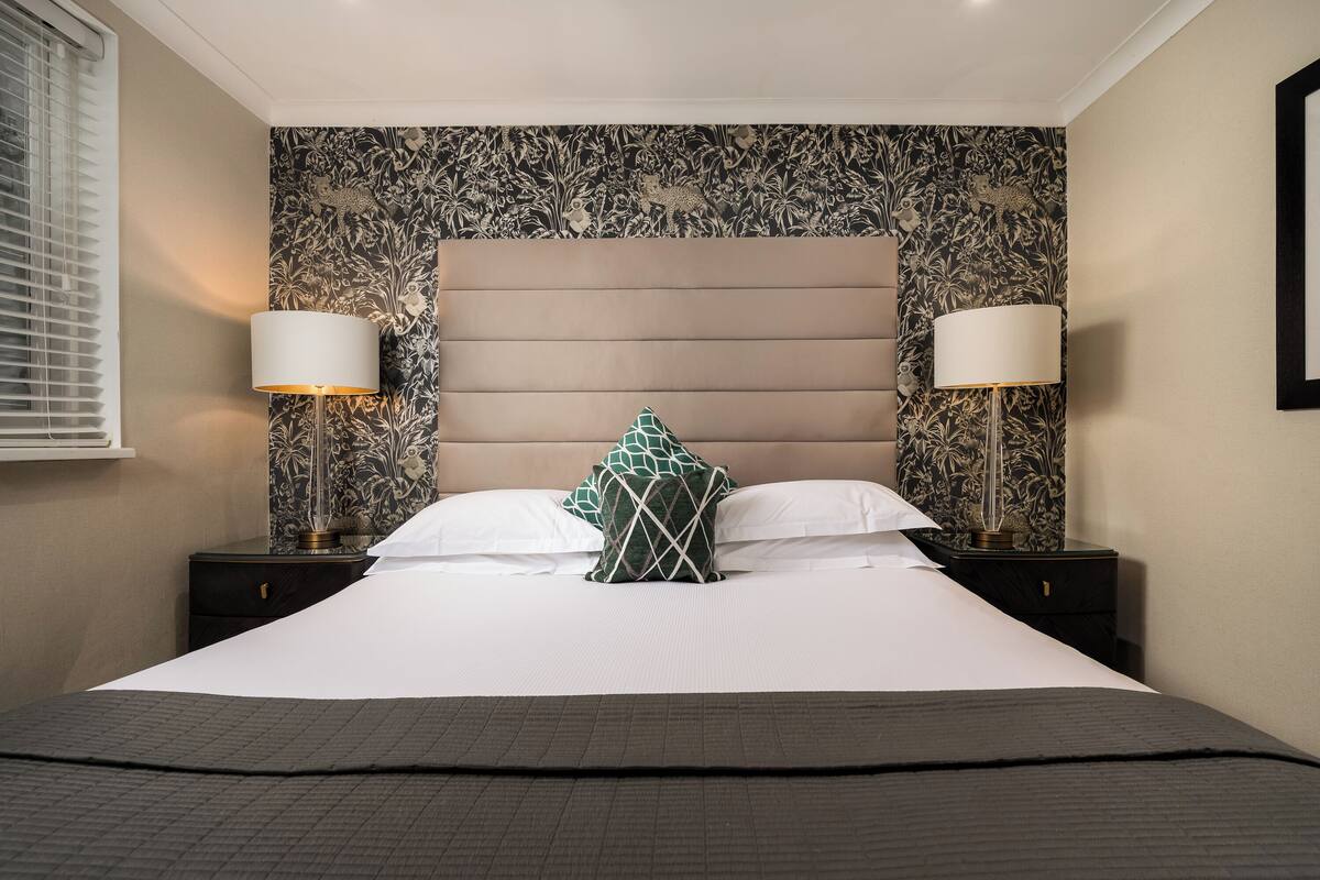 A neatly made bed with a headboard, two bedside lamps, and a patterned wallpaper.