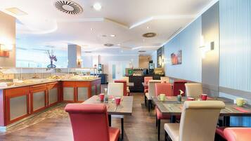 Ein Hotelrestaurant mit modernem Interieur, das eine zentrale Bar, mehrere Speisetische mit Stühlen und eine im Hintergrund sichtbare Küche bietet.