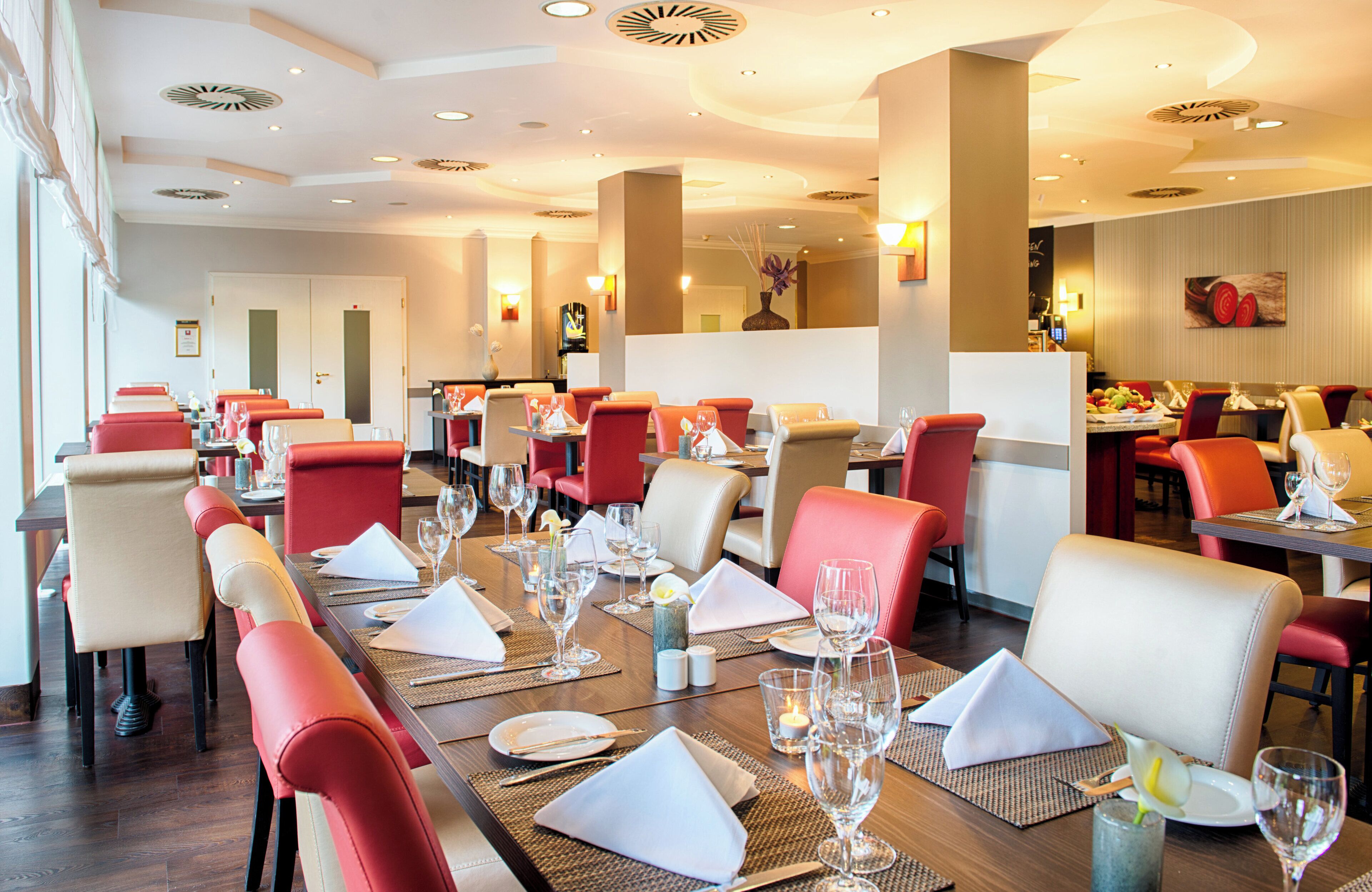 Ein Restaurant mit mehreren Tischen, roten und beigen Stühlen und einem modernen Innenraum.
