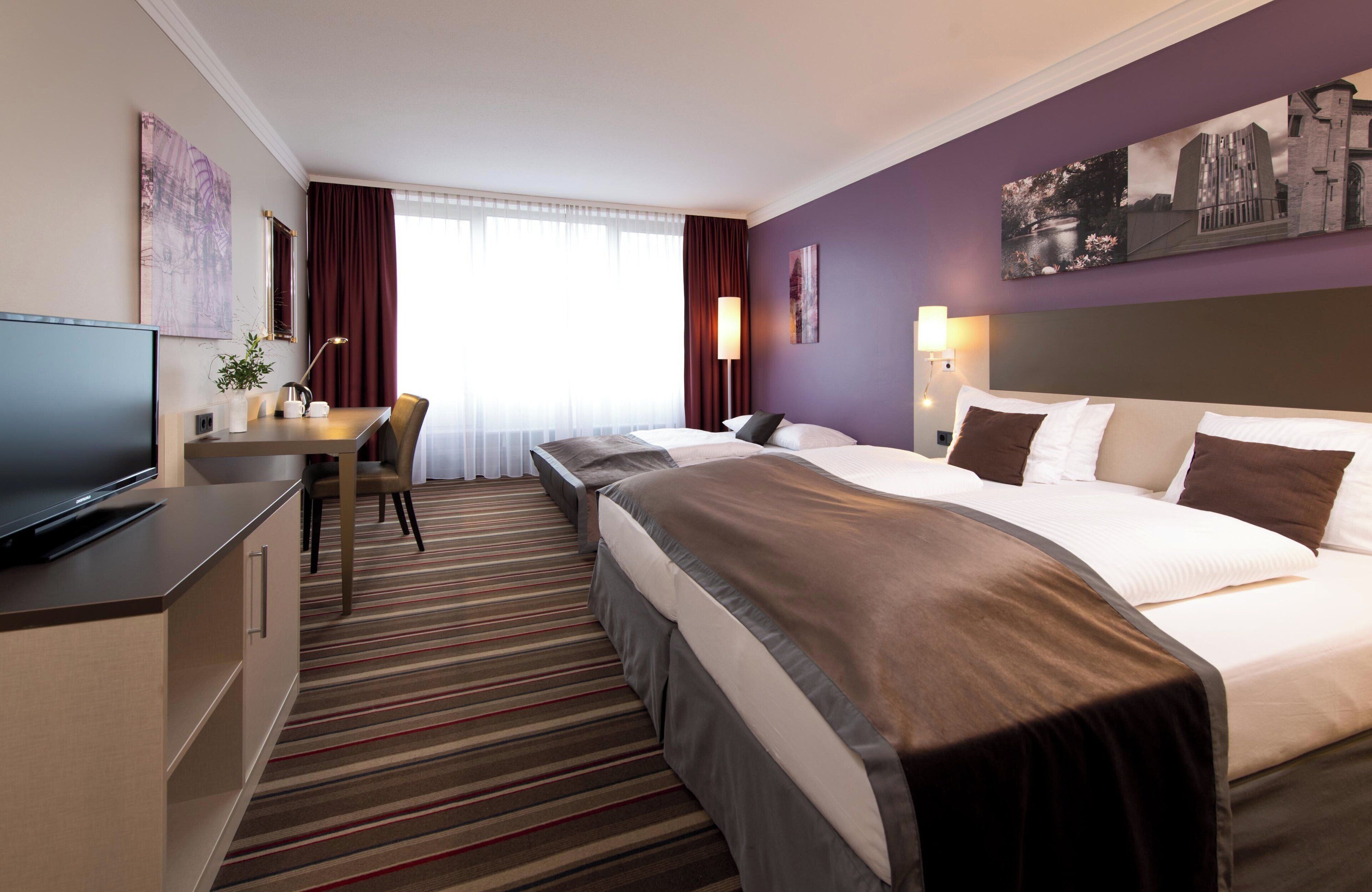 Ein Hotelzimmer mit Bett, Schreibtisch mit Fernseher, Stuhl und einem Fenster mit Vorhängen.