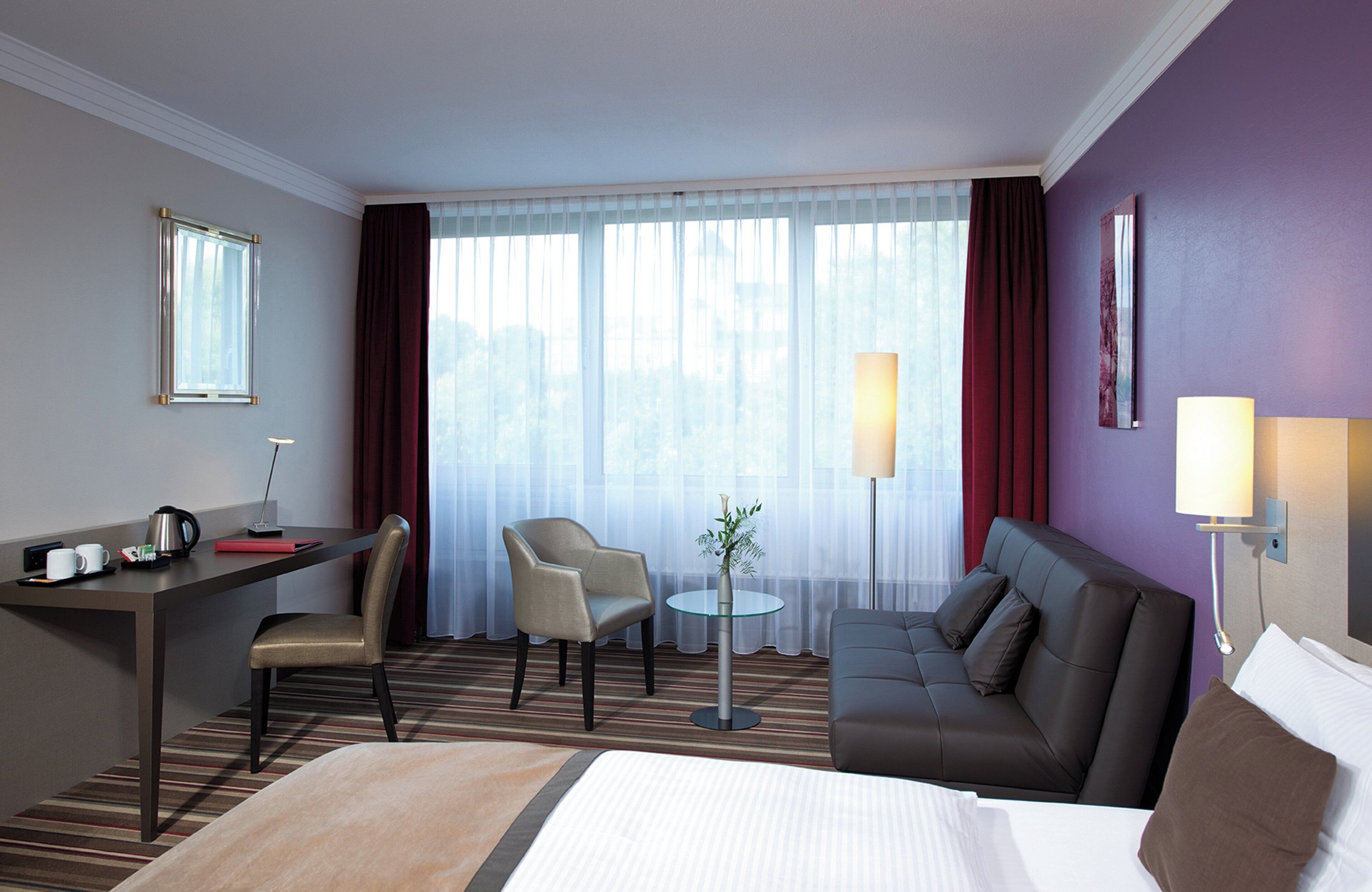 Habitación de hotel con cama, escritorio, sillón, sofá, lámpara y vistas a la ciudad a través de amplias ventanas.