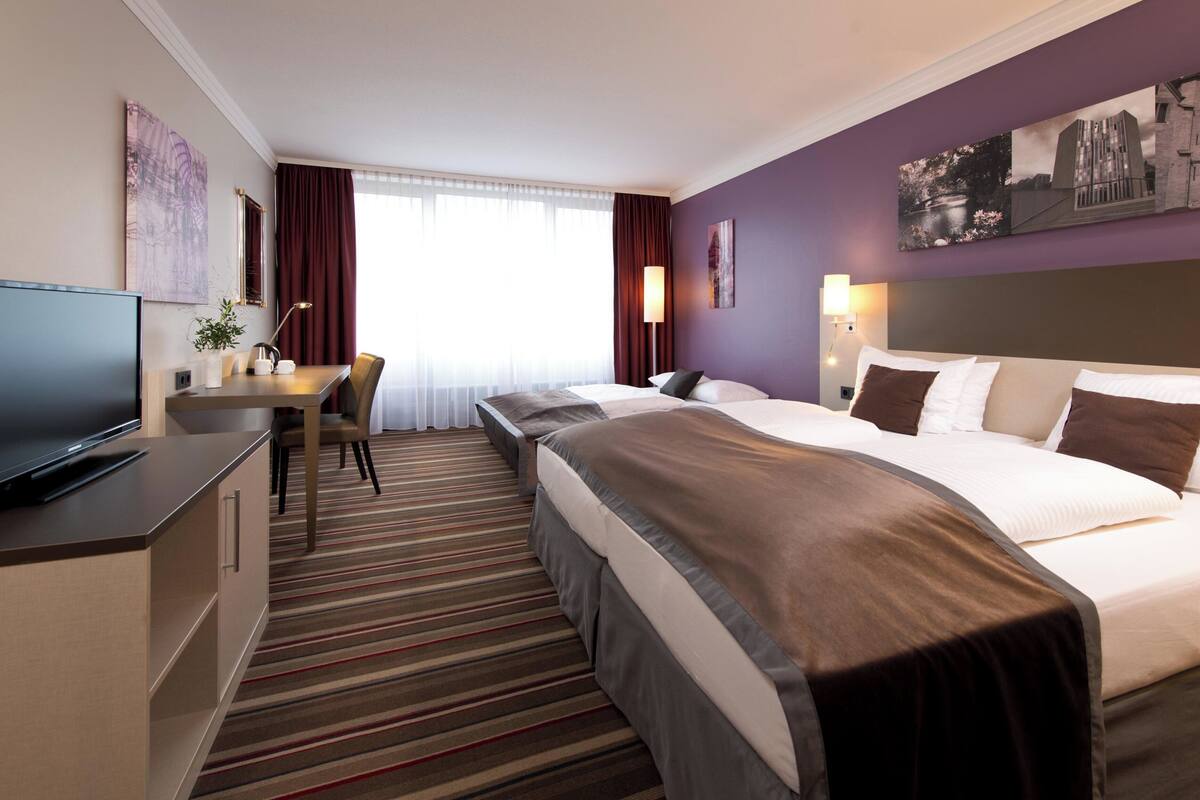 Una habitación de hotel con cama, escritorio con televisor, silla y ventana con cortinas.