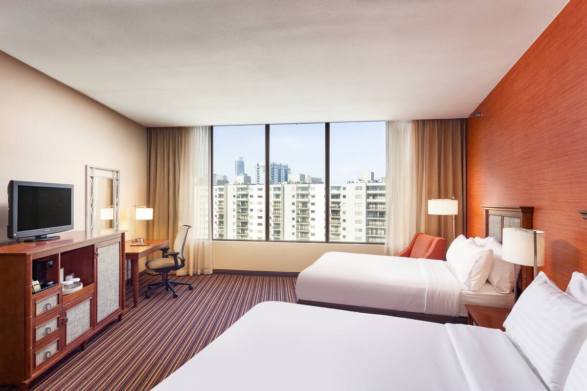A hotel room with two beds, a desk, a TV, and a view of the cityscape.