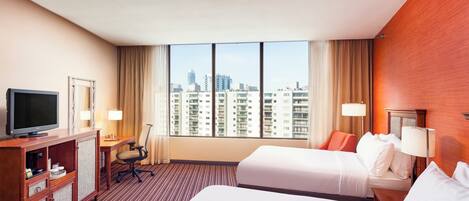 A hotel room with two beds, a desk, a TV, and a view of the cityscape.
