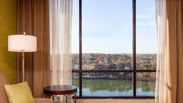 A hotel room with a large window offering a view of a lake and autumn foliage.