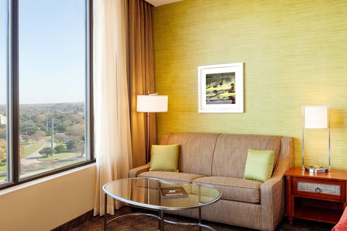 A hotel room with a sofa, a glass coffee table, a lamp, and a framed picture on the wall.