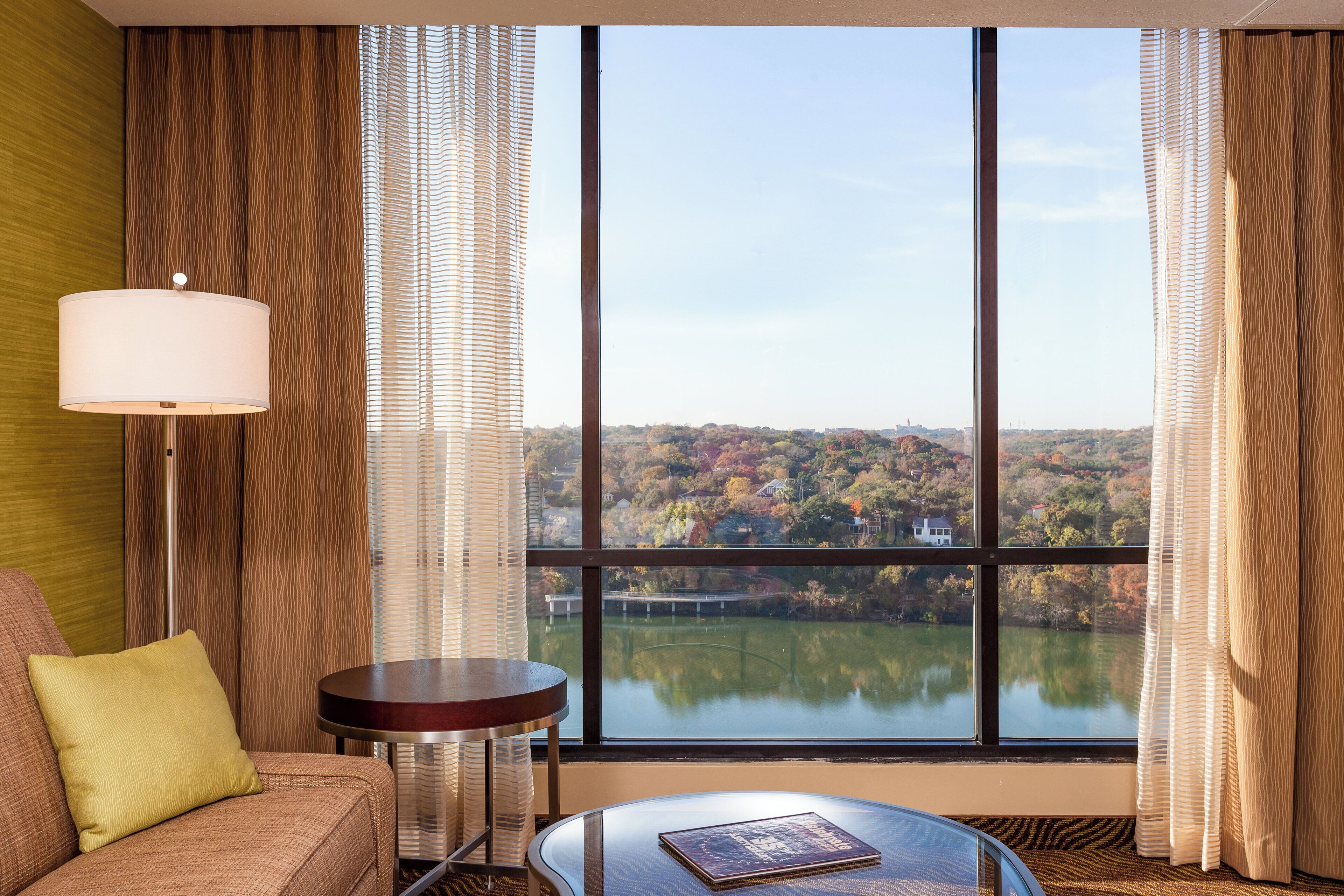Habitación de hotel con un ventanal grande, un sofá, una mesa redonda y una lámpara.