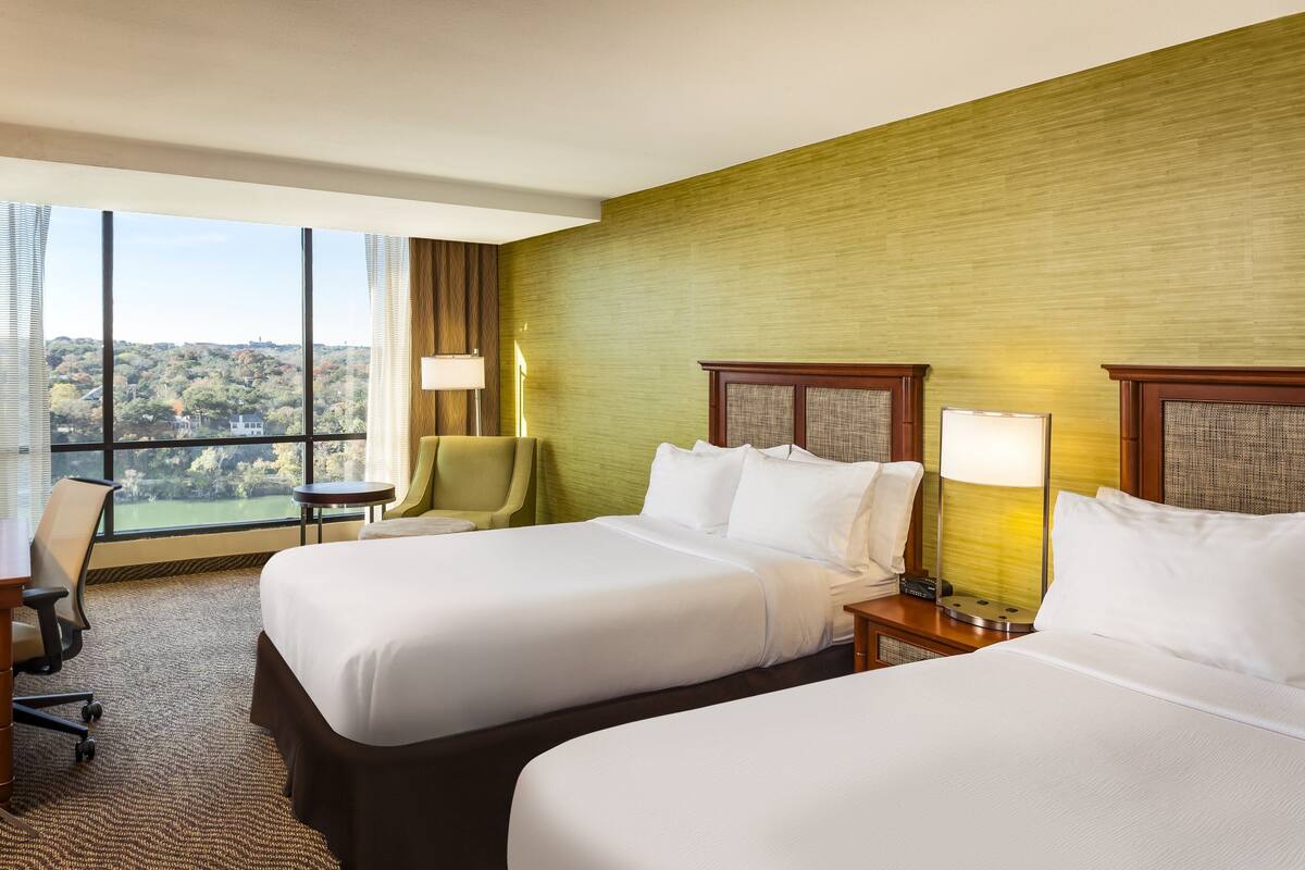 A hotel room with two beds, a desk, a chair, and a large window offering a view of the outdoors.