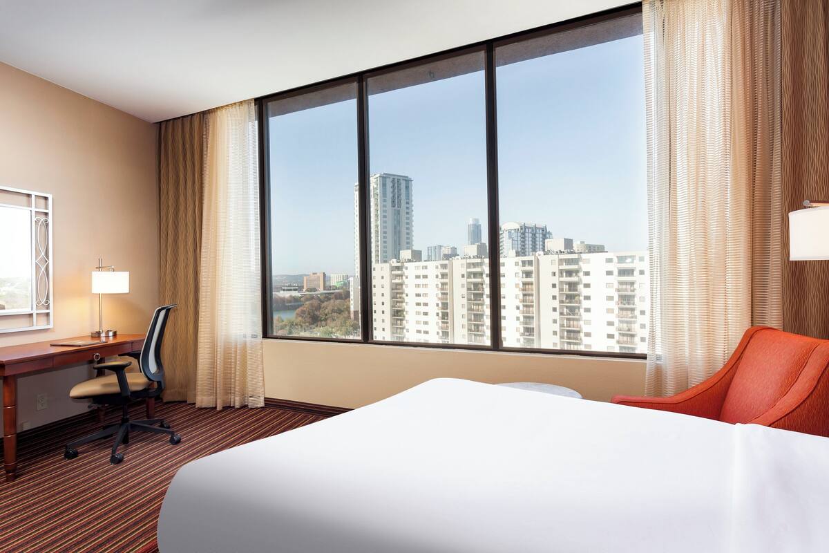 A hotel room with a large window, a bed, a desk with a chair, and a view of the cityscape.