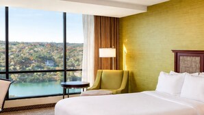 A hotel room with a large bed, a green armchair, a small table, and a view of a lake and hills.