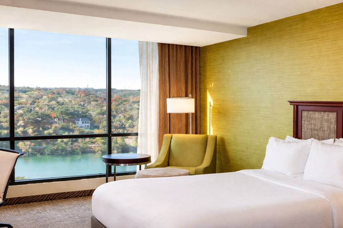 A hotel room with a large bed, a green armchair, a small table, and a view of a lake and hills.