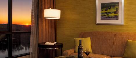 A hotel room with a glass table, a sofa, a lamp, a bottle of wine, and a plate of cheese and fruit.