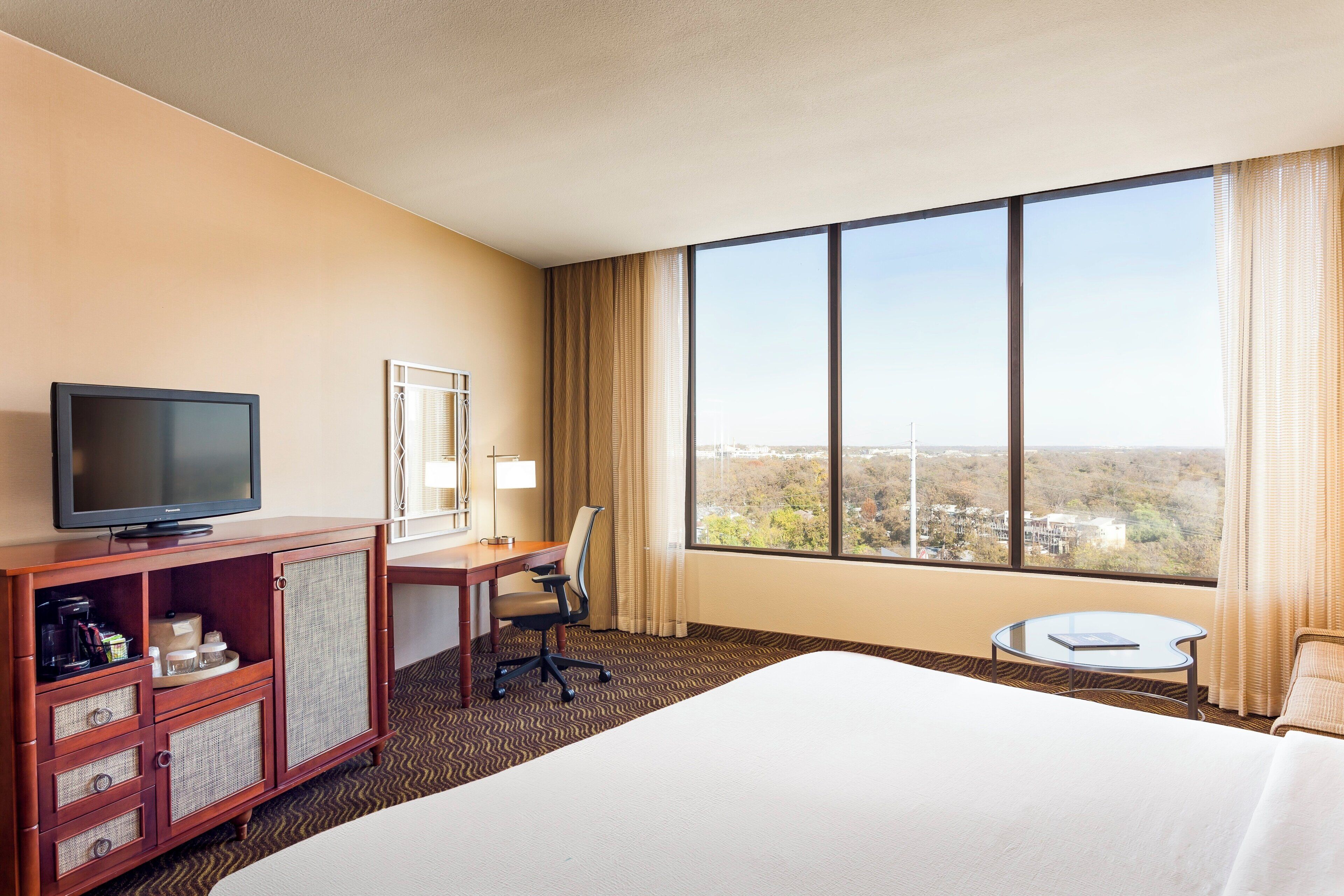 A hotel room with a large bed, a desk with a chair, a television, a small table, and a window with a view of buildings.