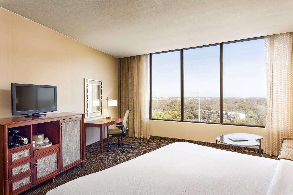 A hotel room with a bed, a desk with a chair, a television, a small table, and a large window with a view.